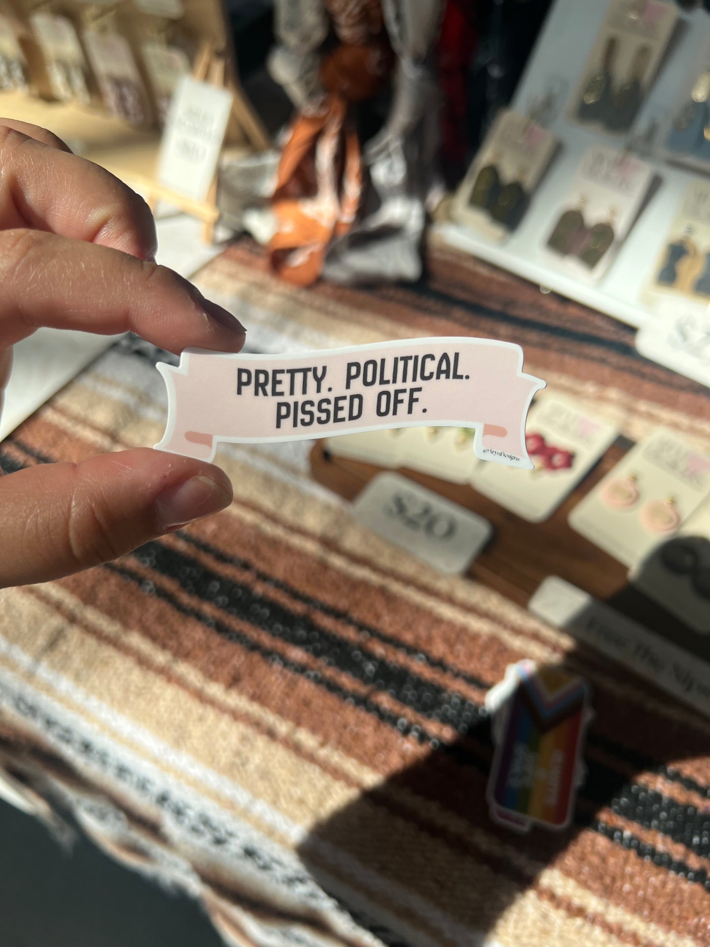 Hand holding a small card with text 'Pretty. Political. Pissed Off.' against a blurred background.
